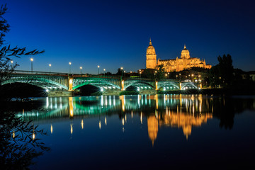 Fototapeta na wymiar Night view of the Cathedral of Salamanca with Enrique Esteban bridge lit foreground and reflections in the Tormes river, Salamanca, Spain