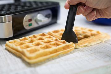 Mit einem schwarzen Spatel werden auf einer weissen Holzplatte zwei Waffeln geteilt. Im Hintergrund...