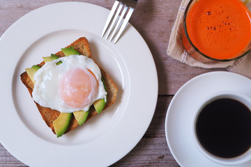 Top view of a breakfast of poached egg and avocado on whole wheat toast with coffee and carrot juice.