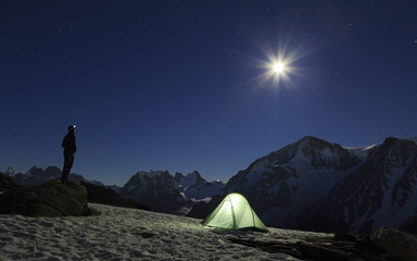 Hiker beside his tent, looking at the moon rising above the mountains in Wallis, Switserland.