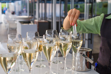 The waiter throws a ice peaces in wine glasses using tongs, Gray table covered with half full glasses. Glasses of white wine for party or wedding.