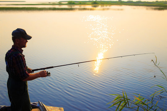 Old Fisherman Throws A Fishing Rod Spinning. The Old Man Is Fishing From A Boat On The River At Sunset