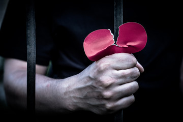 Teenager standing behind iron bars. One  hand holding a heart made out of paper.

