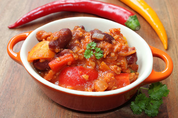 chili con carne mit chillies, frischem Gemüse, Petersilie und Gewürzen in einer kleinen orangenen Schüssel mit Löffel auf einem braunen Holztisch