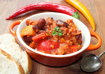 chili con carne mit chillies, frischem Gemüse, Petersilie und Gewürzen in einer kleinen orangenen Schüssel mit Löffel und Brot auf einem braunen Holztisch