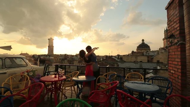 Young loving couple softly kissing on the rooftop cafe with ancient city view while raining. Romantic sunset with sky in clouds on the background