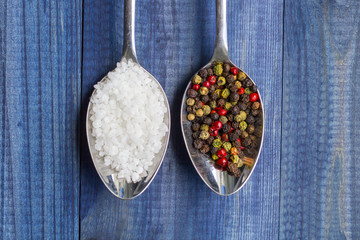 Salt and pepper. Two spoons with salt crystals and color peppercorns on blue wooden background.