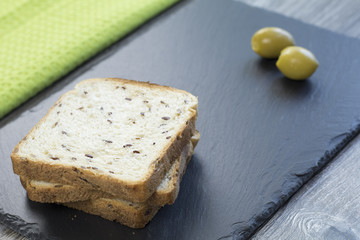 Fresh whole grain toast with linseed and two green olives on a black stone tray and green cloth in the background.