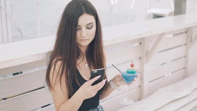 Young girl using phone with goblet of cocktail in hand on the beach bench in 4K