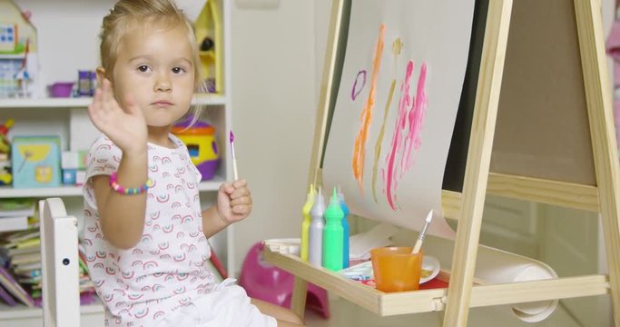 Pretty little girl artist sitting at an easel painting a colorful abstract picture with watercolors pausing to look earnestly at the camera