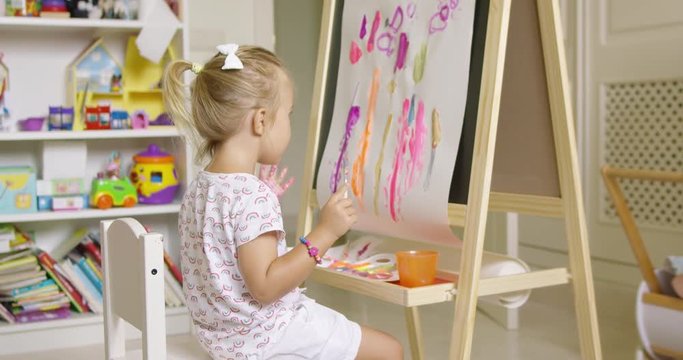 Little girl painting with her hand applying a palm print to a colorful abstract picture on an easel at home with brushes and an artists palette in front of her.