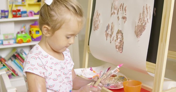 Cute little girl choosing a paint brush from a selection clasped in her hand as she sits at an easel painting in her playroom
