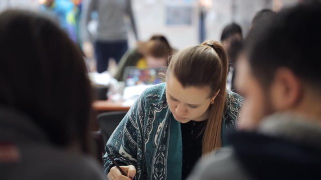 A young woman graduate student uses a washing elastic band and pencil, in middle of people around her.