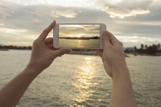 Close up of woman's hand holding smart phone, mobile, smart phone over blurred beautiful sea with sunset moment to take a photo of the sea,selective focus,filtered image
