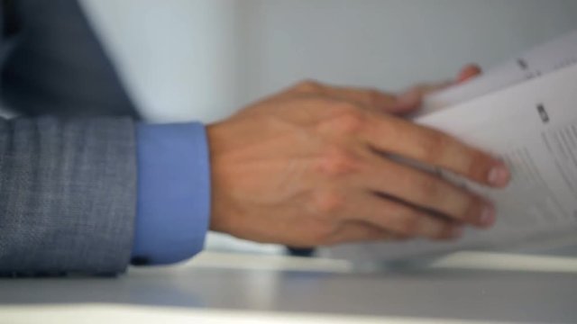 Male notary in suit jacket and light blue shirt holds a document and reads them while sitting at table, closeup.