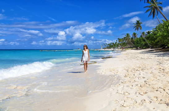 Young woman walking along white sand tropical beach. Beautiful girl in white dress on the beach. Travel and vacation. Freedom concept.
