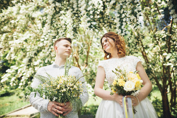 Stylish couple of happy newlyweds walking in the park on their wedding day with bouquet