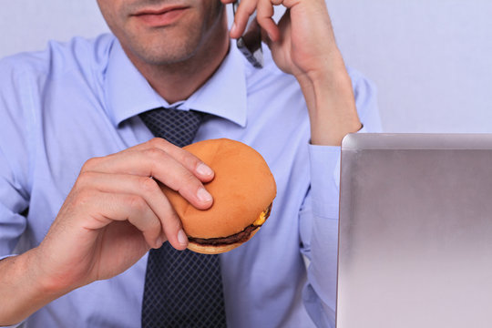 Overworked Businessman Eating Hamburger / Junk Food For Diner In An Office. Unhealthy Eating Leads To Chronic Gastritis Or Ulcer