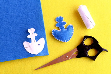 Home made anchor. Blue felt anchor is sewn with white thread, needle, paper pattern pinned on a flat piece of blue felt, scissors on a yellow background. Sewing instruction for children. Step