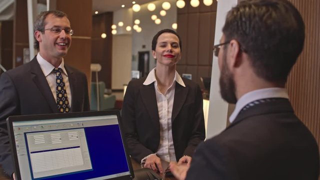 Business Man And Woman Going To The Reception In The Hotel To Check In. They Giving Documents And Credit Card To Man At The Register And Talking To Him With Smile