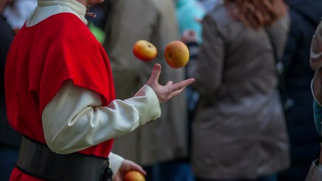 A young court jester is juggling with apples in among the visitors who are walking around and trying different foods. They came to see a battle of knights. Close-up shot.
