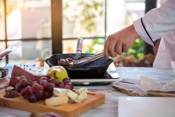Tongs touching meat on pan. Wooden board and small cooker. Fried beef with spices. Dish ready to be served.