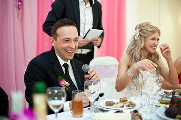 Laughing groom holds a microphone sitting behind a bride