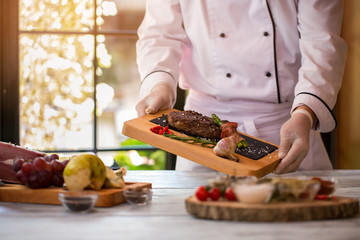 Cook holds board with meat. Garlic and chili peppers. Ribeye steak served with rosemary. Tasty lunch for a gourmet.