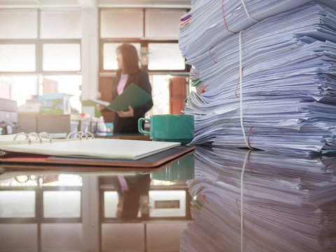 Pile of unfinished documents on office desk with businesswoman background