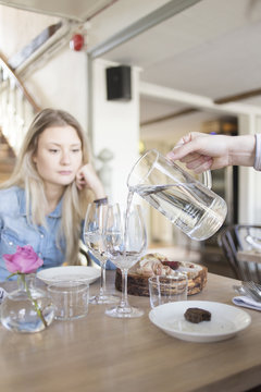 Cropped image of hand pouring water in wineglass while woman sitting at restaurant table