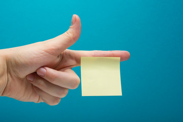 Sticky note, finger up of thumb, yellow reminder on blue background.