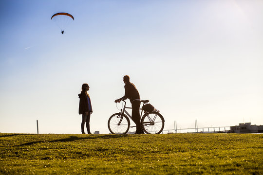 Couple with bicycle watching paraglider in mid-air from grassy landscape