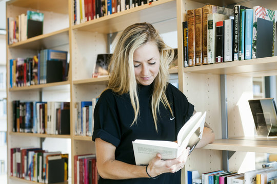 Young woman reading book while leaning on bookshelf in college library