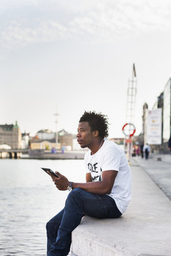 Thoughtful male student holding digital tablet while sitting on retaining wall by river
