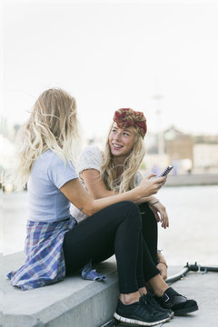 Female college students with mobile phone while sitting on retaining wall by river