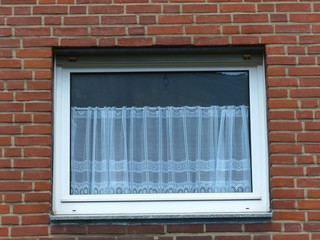 Fenster mit spießiger Gardine in einer Fassade aus braunem Backstein bei Sonnenschein im...