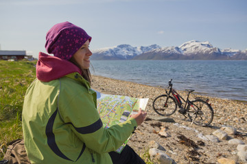 Eine junge Frau schaut in die Karte, dabei sitzt sie am Strand/ Norwegen. Fahrrad im Hintergrund...