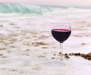 glass with red wine in a white sea foam on the beach. sea pebbles in the water. In the background, emerald wave rolls ashore. Natural beautiful backdrop and sea glass
