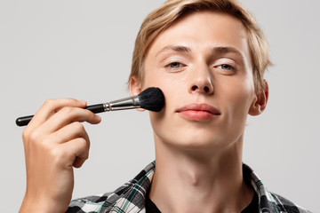 Funny handsome blond young man wearing casual plaid shirt with make-up brush looking in camera over grey background, copy space