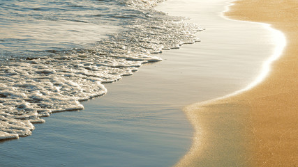 Close up of surf with soft sea waves rushing on sandy beach on sunset with sun reflection. Enjoying summer vacation on tropical country. Nature background