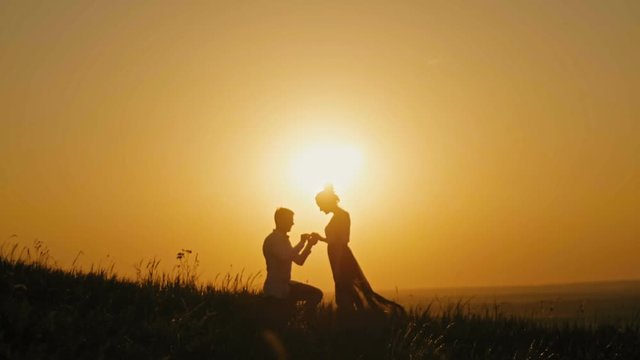 Romantic Silhouette of Man Getting Down on his Knee and Proposing to Woman on high hill - Couple Gets Engaged at Sunset - Man Putting Ring on Girl's Finger, slow motion