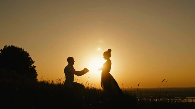 Romantic Silhouette of Man Getting Down on his Knee and Proposing to Woman on high hill - Couple Gets Engaged at Sunset - Man Putting Ring on Girl's Finger