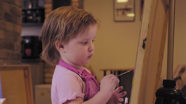 The little girl in a pink apron paints on a canvas in the studio thinks, smiles and draws on.