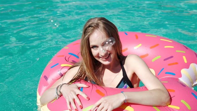 Young hipster millennial girl in sprinkled donut float at pool, festival, party, hotel, beach, event smiling with sunglasses. 20s. 1080p Slow Motion