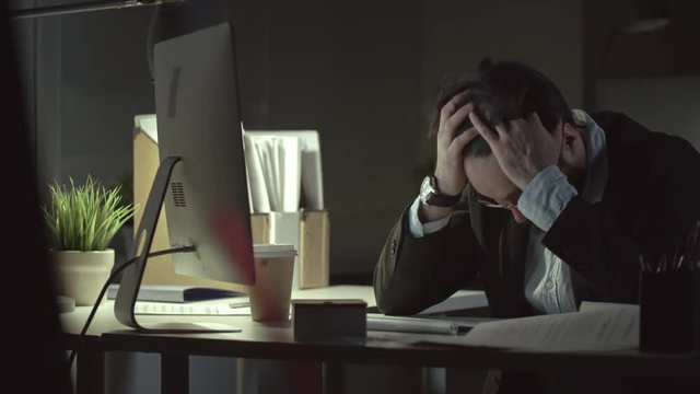 Desperate businessman sitting alone at the desk in the dark office at night and holding head in hands
