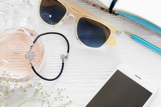 Fashionable woman workplace with female accessories. Smartphone with blank screen with sunglasses, perfume, bracelet, pen and notebook on white wooden table, mockup