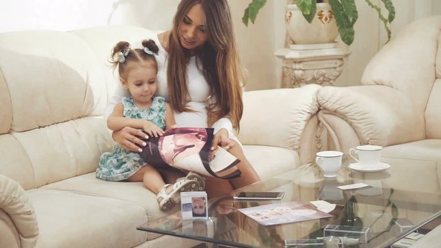 Pretty woman with little girl sitting in comfortable room, looking on magazine