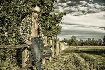 Portrait of sexy farmer or cowboy in hat looking at camera while leaning on wooden fence in...