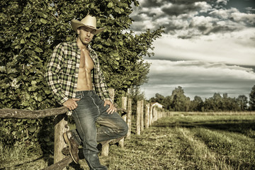 Portrait of sexy farmer or cowboy in hat looking at camera while leaning on wooden fence in...