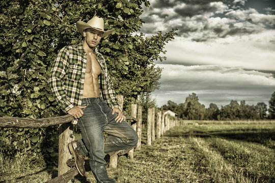 Portrait of sexy farmer or cowboy in hat looking at camera while leaning on wooden fence in countryside